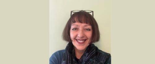 Carys has short brown hair, wears glasses on her head and a blue jumper with a blue scarf