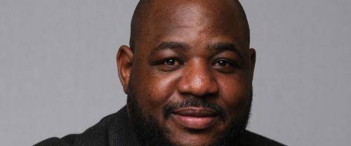 Carlton wears a clerical collar with a black shirt and jacket