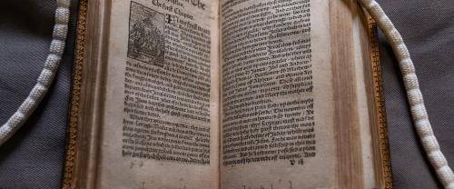 Tyndale Bible open at "The Acts of the Apostles". Long white "snake" weights are visible holding the top right and left corners of paper open. 